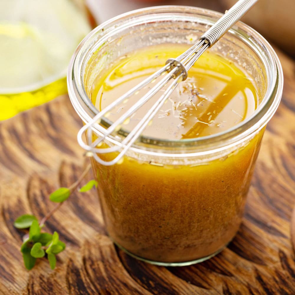 a jar of lemon garlic marinade with a silver whisk on top of the jar placed on a brown wooden chopping board