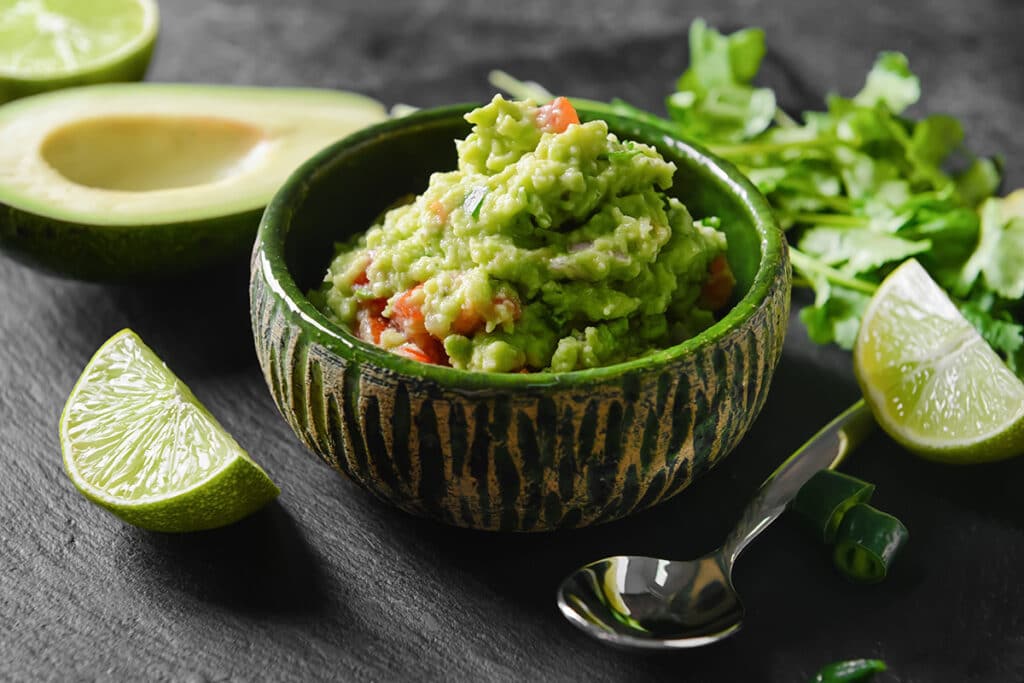 guacamole in a green bowl surrounded by avocado, lime, silver spoon, and cilantro