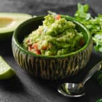 guacamole in a green bowl surrounded by avocado, lime, silver spoon, and cilantro