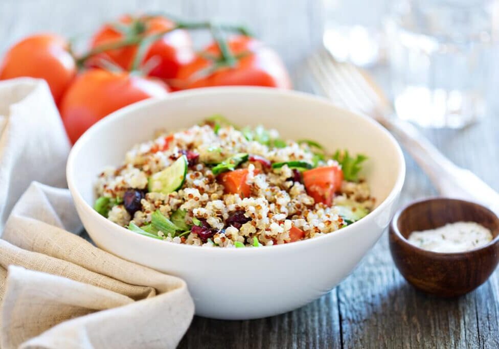 mediterranean quinoa salad in a white bowl beside a kitchen cloth, four pieces of red tomatoes, a fork, and bowl of sauce no a brown wooden table