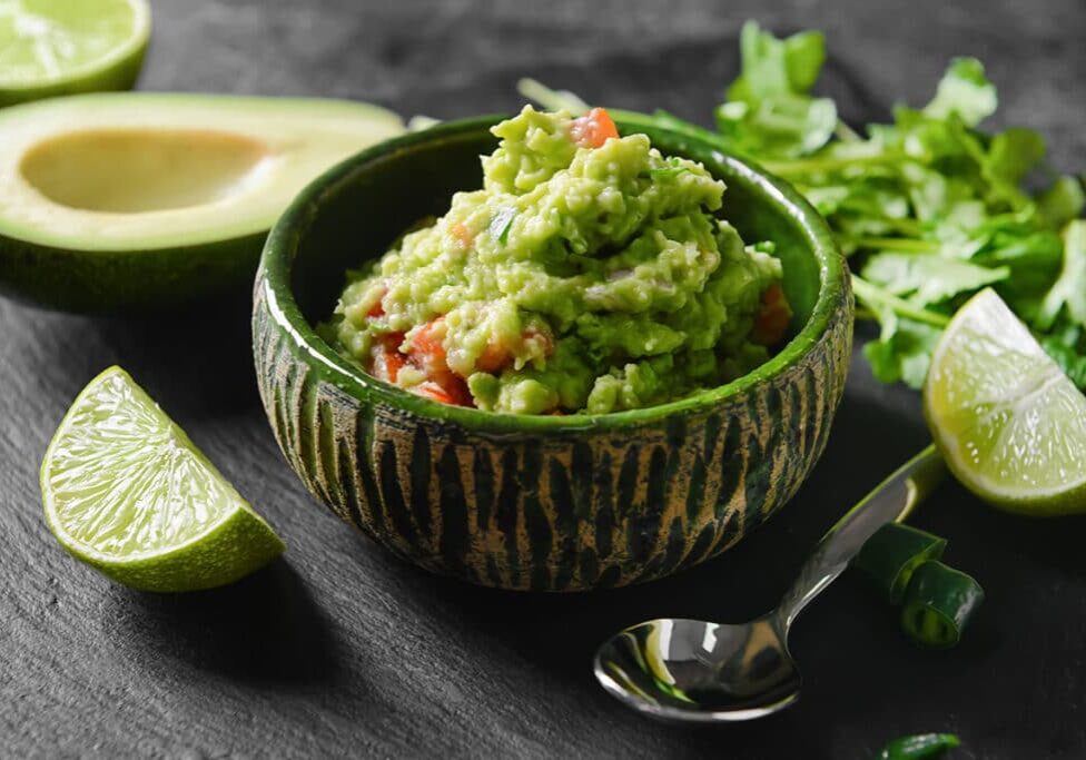 guacamole in a green bowl surrounded by avocado, lime, silver spoon, and cilantro