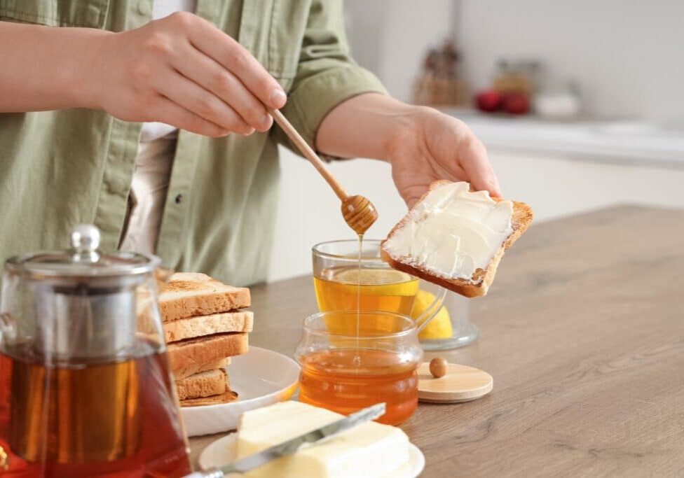 Young,Woman,Adding,Sweet,Honey,Onto,Toasted,Bread,In,Kitchen